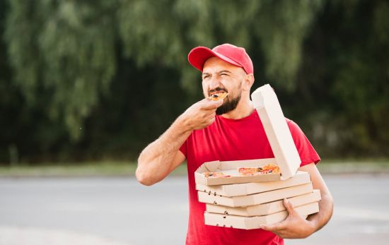 Un repartidor come un trozo de pizza rápido y de pie/ FREEPIK Un repartidor come un trozo de pizza rápido y de pie/ FREEPIK