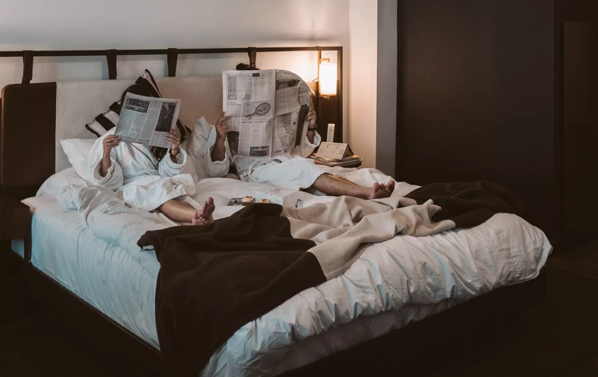 Dos huéspedes leen la prensa en una cama de hotel / PEXELS
